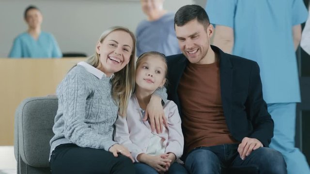 Portrait of the Young Happy Family in the Hospital. Handsome Father, Beautiful Mother and Cute Little Daughter Sitting in the Lobby of the Medical Facility, Smiling. 