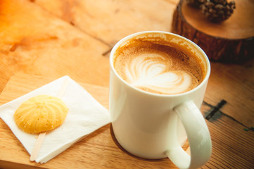 Hot latte art in heart shape in white coffee cup and cookie on wooden background