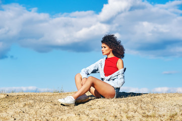 Young woman sitting on sand on sky background . Girl in shorts and shirt in nature