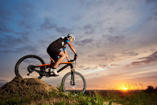 View of energetic and active man in helmet and sportswear riding orange bike in the evening. Sporty and energetic athlete sitting and rolling down hill at sunset. Recreative fresh air activities.