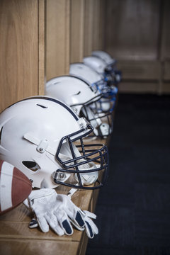 Close up view of a row of American football helmets sitting in a locker room before an football game. Gloves and a football also sitting on the bench in the locker room