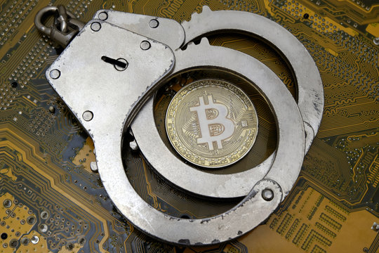 The danger of electronic fraud, hacker attacks or violation of the law in the crypto-currency area. Bitcoin lies under police steel cuffs against the background of a computer electronic motherboard.