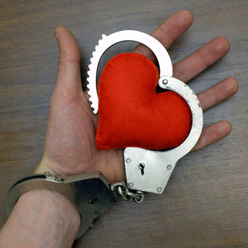 The hand of the man is handcuffed, holding a red soft heart on a brown background. A square picture. The concept of love and the law, heart disease.