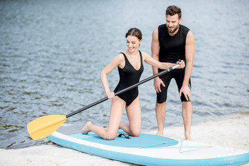 Male coach teaching young woman to surf on the standup paddleboard with oar standing on the beach
