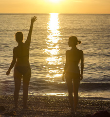 two girls standing on sandy beach near mediterranean sea at sunset