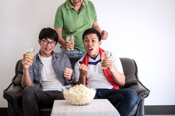Young Happy Asian Men family or football fans watching soccer match on tv and cheering football team, celebrating with drink beer and eat popcorn at home, sports and entertainment concept