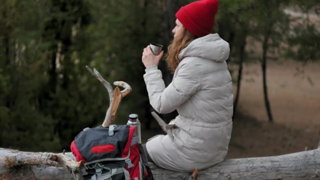 An attractive young woman in a red hat walks through the forest in early spring with a large tourist backpack, drinks tea from a thermos