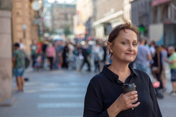 Nice portrait of a middle aged older woman walking in city. Outdoor headshot of 45 50 year old relaxed woman on lunch break drinking coffee. Urban background. Street style shot.