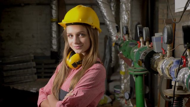 Young cute girl builder is standing near pipes, smiling, crossing arms, watching at camera, building conception