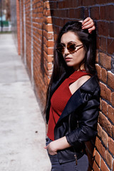 model in sunglasses, black leather jacket, jeans. Posing near a red brick wall.