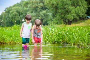 Two little brothers playing with paper boats by a river on warm and sunny summer day.