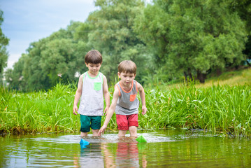 Two little brothers playing with paper boats by a river on warm and sunny summer day.