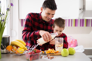Gorgeous little boy and his loving father pouring milk together in fruit mixture in blender bowl.