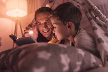 Before sleep. Nice pleasant caring father holding a flash light and smiling while reading a fairy...