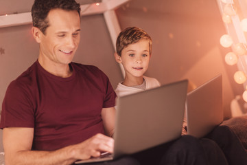 From home. Joyful happy smart man sitting together with his son and looking at the laptop screen while working on the laptop