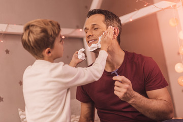 Interesting activity. Delighted pleasant nice boy looking at his father and putting foam on his cheeks while helping him to shave