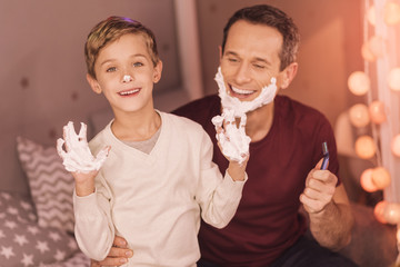 Shaving foam. Happy positive delighted boy looking at you and smiling while showing his hands in the shaving foam