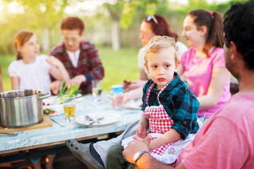 Little boy with adorable little apron sitting on his father lap and looking at the distance while the rest of the family is sitting eating at the picnic table and eating.