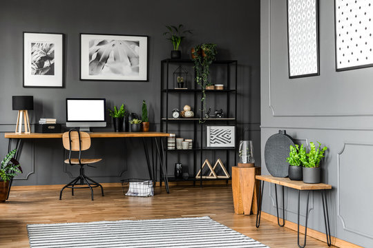 Black rack with decorations standing next to a wooden chair and desk with plants, lamp and a computer in home office interior. Real photo