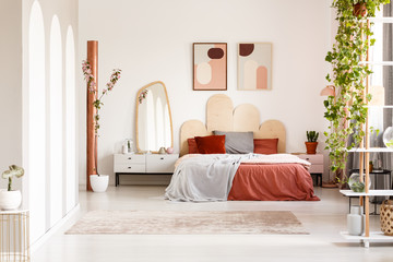 Mirror on cabinet next to orange bed under posters in bright bedroom interior with plants. Real...