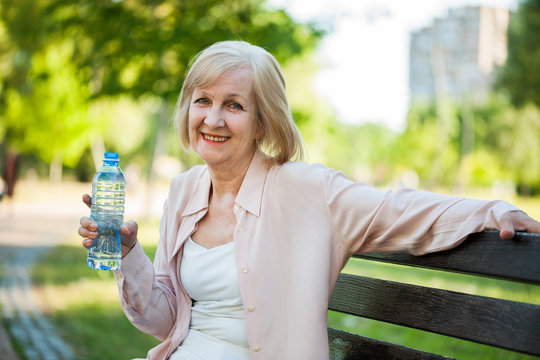 Adult woman is sitting in park and drinking water. 