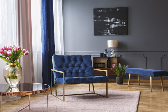Real photo of a blue, wide chair standing on a rug in a spacious living room interior with grey walls and wooden floor next to a shelf and a table