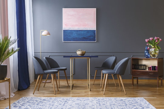 Real Photo Of Four Chairs Standing Around A Table In A Spacious Dining Room Interior Next To A Patterned Rug, Shelf, Lamp And Grey Wall With A Painting