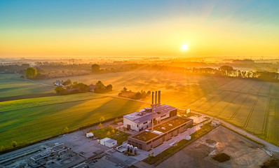 Fototapeta premium Zur goldenen Stunde am frühen Morgen, eine Fabrik vor einem Feld im Hintergrund bei gelb glühender Sonne vor einem See von oben als Luftaufnahme einer Drohne