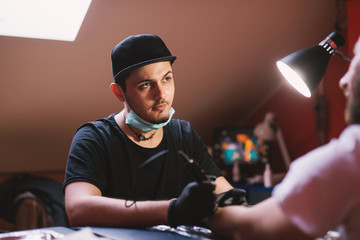 Young focused tattoo artist is inking customers arm carefully in his shop.