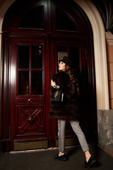 Glamorous woman with long wavy brunette hair dressed in gray trousers and luxury fur coat, holding elegant handbag, opening antique wooden door to enter building. Gorgeous female model in outerwear.