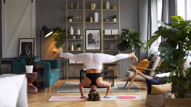 Attractive Caucasian woman in her thirties does yoga poses headstand and bend in the morning in stylish sunny spacious living room in elegant Scandinavian style decorated with sophisticated decor