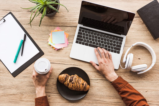 cropped image of man working at table with laptop, headphones, textbook, clipboard, post it, potted plant, coffee cup and croissant