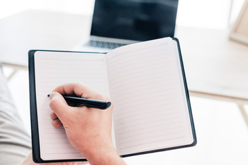 Fototapeta premium cropped image of man writing in textbook at table with laptop