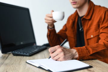 partial view of man drinking coffee and writing in textbook at table with computer and computer keyboard