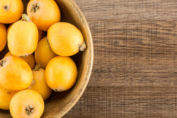 Fresh orange Japanese loquats in a wooden bowl flatlay isolated on brown wood background.
