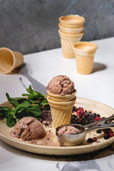 Homemade chocolate raspberry ice cream in small waffle cup served with frozen berries, fresh mint, chopped dark chocolate and metal spoon in ceramic plate on white marble kitchen table.