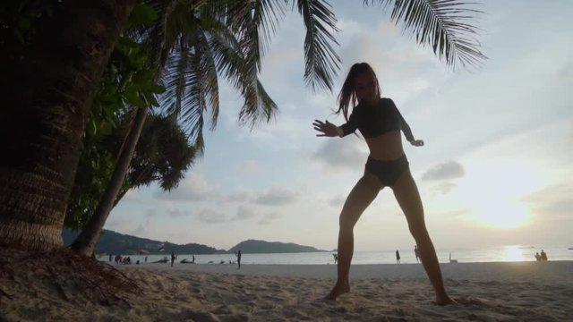 Bottom view of beautiful woman in black swimwear dancing on the sandy beach during sunset over palm tree and sky background - video in slow motion