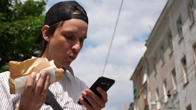 Young man stay in city public place use mobile phone and eat a big sandwich