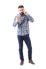 Bearded hipster man twirling mustache using mobile phone reflection as mirror. Full body isolated on white background. 