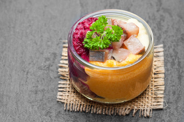 Herring  salad with colorful vegetables in a small glass jar on black stone background
