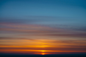 Red sun circle rises out from behind dark horizon on background varicolored clouds of warmly shades. Beautiful background of dawn on picturesque cloud sky. Sun in center.