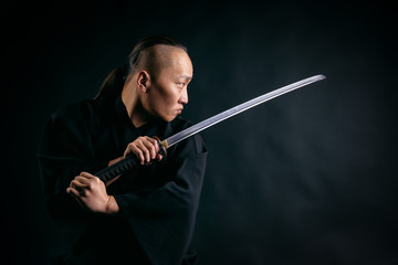Asian man with a sword in his hands in a black robe against a black background