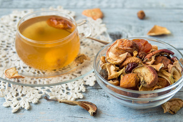 A mixture of dried fruits in a small round glass bowl, next to a mug with compote on an old table.