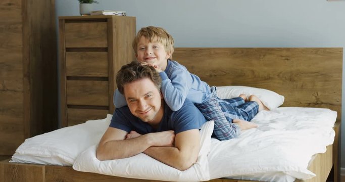 Portrait shot of the smiled Caucasian father and son lying in the pajamas on the bed in the morning, small boy lying on his dad's back. Indoor