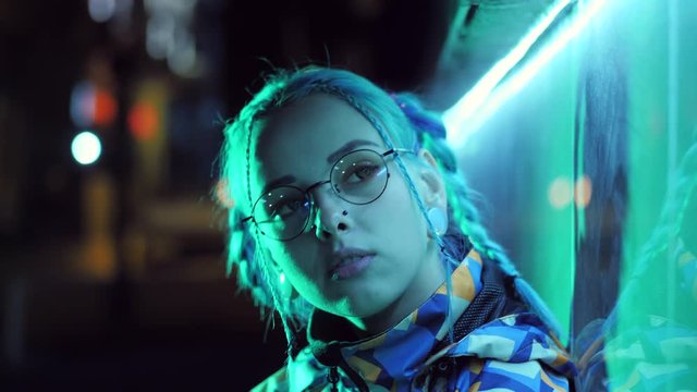 Young pretty girl with unusual hairstyle near glowing turquoise neon light of the city at night. Dyed blue hair in braids. Pensive sad hipster teenager in glasses.