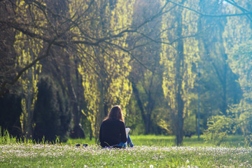 Lonely girl reading a book in a park, she's sitting on a flowered garden (back view of young woman...