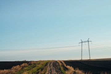 Atmospheric landscape with power lines in green field with dirt road under blue sky. Background image of electric pillars with copy space. Wires of high voltage above ground. Electricity industry.