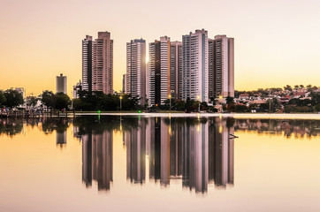 Fototapeta na wymiar Growing city with few tall buildings reflected on the water of a lake, sunset hour - golden hour. Campo Grande MS, Brasil