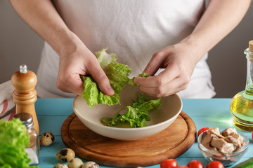 Chef tears the lettuce leaves into ceramic bowl on kitchen table with fresh ingredients.