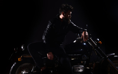 Plakat Man with beard, biker in leather jacket lean on motor bike in darkness, black background. Biker culture concept. Macho, brutal biker in leather jacket stand near motorcycle at night time, copy space.